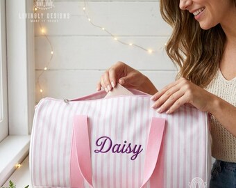 May include: A duffel bag featuring a white and light pink vertical striped pattern with two pink fabric handles. The name Daisy is embroidered in purple script lettering on the front center of the bag. The bag has a zipper closure at the top.