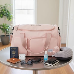 K&ouml;nnte beinhalten: Eine rosa Reisetasche mit dem Namen "Emilia" aufgestickt, umgeben von Reisezubeh&ouml;r. Dazu geh&ouml;ren ein Reisepass, eine Sonnenbrille, ein Reisekissen und eine kleine Flasche H&auml;ndedesinfektionsmittel.
