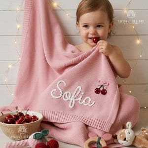 Puede incluir: Una manta rosa tejida para bebé con el nombre "Sofia" bordado en blanco. La manta presenta un diseño de cerezas y está sobre un bebé. Un cuenco de cerezas y un juguete también están en la imagen.