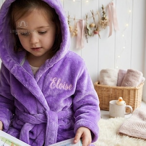 May include: A plush purple hooded bathrobe featuring long sleeves, a tie belt at the waist, and the name Eloise embroidered in light purple thread on the left chest area. The fabric has a soft, fuzzy texture throughout the entire garment.