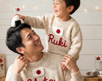May include: Two matching cream-colored knit sweaters featuring a red embroidered rose emblem above the names Ruki and Yuto in red cursive script. The sweaters have a textured knit pattern and a crew neckline, showcasing a coordinated design.