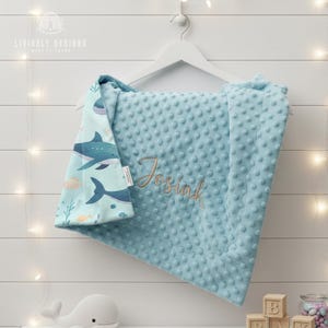 May include: A baby blanket in light blue with a minky dot texture and a shark print. The blanket is personalised with the name "Josiah" in gold thread. The blanket is hanging on a white hanger.