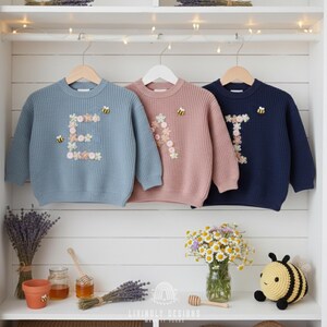 May include: Three children's sweaters in blue, pink, and navy blue, each with a floral initial and a bee applique. The sweaters are hanging on wooden hangers against a white background. The bottom shelf displays lavender, honey, and a crocheted bee.
