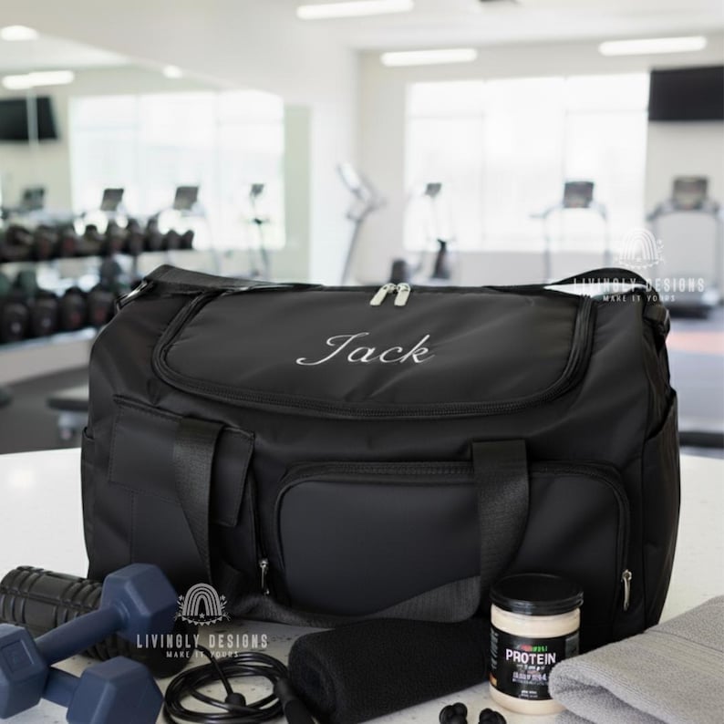 K&ouml;nnte beinhalten: Schwarze Sporttasche mit dem Namen "Jack" in Wei&szlig; gestickt. Die Tasche hat mehrere Taschen und wird mit Hanteln, einem Springseil, einem Handtuch und einem Proteinpulverbeh&auml;lter gezeigt. Der Hintergrund ist ein Fitnessstudio.