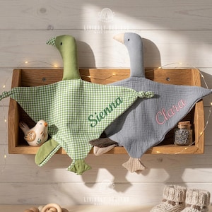Puede incluir: Dos mantas de bebé personalizadas con forma de ganso. Una es de cuadros verdes con "Sienna" bordado en verde, la otra es gris con "Clara" bordado en rosa. Se exhiben en una estantería de madera con un pájaro de cerámica y un tarrito.