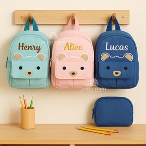 Może przedstawiać: Trzy spersonalizowane plecaki z motywem misia w kolorze morskim, różowym i granatowym, każdy z imieniem: "Henry", "Alice" i "Lucas". Poniżej znajduje się pasujący granatowy piórnik. Na stole znajduje się drewniany pojemnik na ołówki i ołówki.