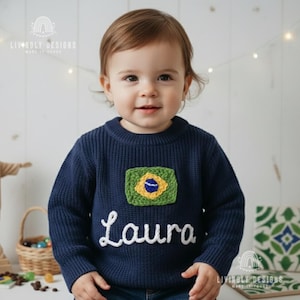 以下が含まれることがあります： ブラジル国旗のパッチと白い筆記体で「Laura」という名前が入った、ネイビーブルーのニットセーター。セーターは小さな子供が着ています。セーターは長袖でクルーネックです。