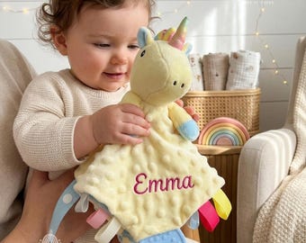 May include: A soft, pale yellow plush unicorn security blanket featuring a textured dimpled fabric, a small embroidered horn, and the name Emma stitched in pink thread. The blanket includes multiple colorful fabric tags and a blue silicone teether.