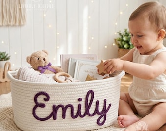 Könnte beinhalten: Ein ovaler Aufbewahrungskorb aus weißem geflochtenem Seil mit dem Namen Emily, der in dickem violettem Garn auf der Vorderseite eingestickt ist. Der Korb enthält einen hellbraunen Teddybären mit violettem Band, einen Holzring und kleine Bücher.