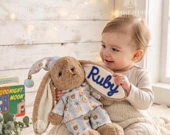 May include: A plush brown rabbit toy wearing light blue pajamas with a teddy bear pattern. The rabbit has a matching nightcap and a cream-colored ear flap with the name Ruby embroidered in dark blue script. The toy has soft fur and buttons.