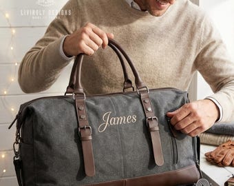 May include: A charcoal gray canvas duffel bag with brown leather trim, featuring two top handles, adjustable buckle straps, and the name James embroidered in a cursive script on the front. The bag includes a side zipper pocket and metal hardware.