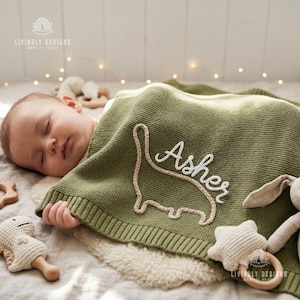 Könnte beinhalten: Ein schlafendes Baby ist mit einer grünen Strickdecke bedeckt. Die Decke zeigt eine Dinosaurier-Umrisslinie und den Namen "Asher" in weißer Schrift. Die Decke ist von Babyspielzeug umgeben.