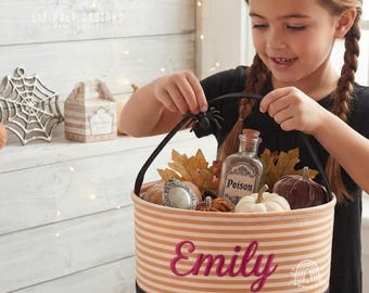 May include: A round fabric basket with horizontal beige and white stripes, featuring the name Emily embroidered in pink script. The basket contains artificial autumn leaves, small pumpkins, and a glass bottle labeled Poison.