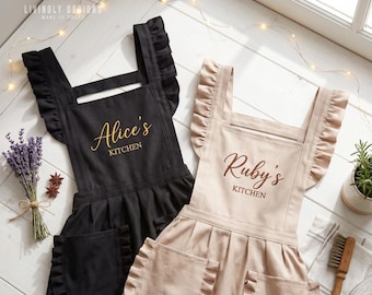 May include: Two aprons with ruffled shoulder straps and two front pockets. One apron is black with gold text reading Alice's KITCHEN, and the other is beige with brown text reading Ruby's KITCHEN. Both feature a pleated skirt design.