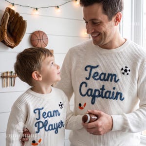 May include: Two cream-colored sweaters with the words "Team Captain" and "Team Player" in blue script. The "Team Captain" sweater has a soccer ball patch. The "Team Player" sweater has a soccer ball patch and a hockey stick patch.