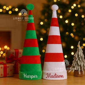 May include: Two personalized Christmas elf hats. One is green and red with the name "Harper", the other is red and white with the name "Madison". Both have a round top and a wide brim. The hats are on a wooden surface with a Christmas tree in the background.