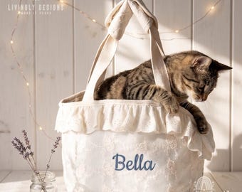 May include: A white fabric tote bag featuring a ruffled eyelet trim along the top edge and the name Bella embroidered in blue script. A brown tabby cat is resting inside the bag, which has two sturdy handles and a delicate floral lace overlay.
