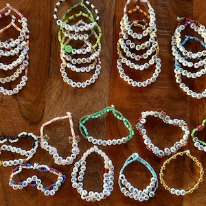 May include: A collection of beaded bracelets with white letter beads spelling out various words and phrases. The bracelets are in various colors, including white, black, red, blue, green, and gold. The bracelets are arranged on a wooden surface.