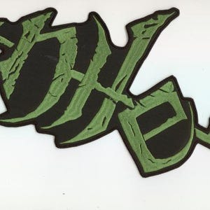 May include: A black patch featuring a green "DK" design. The letters have a cracked, textured appearance, outlined in black. The patch is set against a white background.