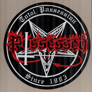 May include: A circular black patch with white and red embroidered details. The patch features the words "Total Possession" and "Since 1983" in white, surrounding a white pentagram with red accents and stylized text. A red arrow points to the bottom.