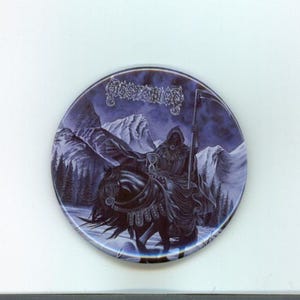 Peut inclure: Bouton rond avec une illustration sombre et gothique. Il présente une figure vêtue d'une robe avec une faux chevauchant un cheval à travers un paysage de montagne enneigé. Le nom du groupe est en haut dans une police stylisée.