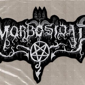 May include: Black embroidered patch with the word "MORBOSIDAD" in white, stylized gothic lettering. The patch features a pentagram, horns, and other intricate white designs on a black background. The patch is likely for a band or related merchandise.