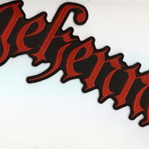 Könnte beinhalten: Ein schwarzer Aufnäher mit dem Wort "Gefenria" in roter gotischer Schrift. Der Aufnäher hat einen dekorativen Rand und ist vor einem weißen Hintergrund platziert.