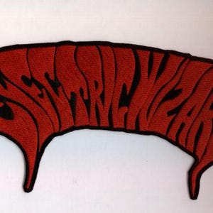 Könnte beinhalten: Ein roter Aufnäher mit dem Wort "STRANGEWEAR" in schwarzer, stilisierter Schrift. Der Aufnäher hat einen schwarzen Rand und eine einzigartige, geschwungene Form mit spitzen Enden, geeignet für Kleidung oder Accessoires.