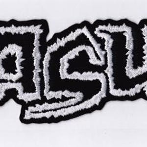 Puede incluir: Un parche bordado en blanco y negro con la palabra "NASUM" en una fuente audaz y estilizada. Las letras son negras con un contorno blanco texturizado, creando un contraste llamativo. El parche tiene una forma ligeramente irregular.