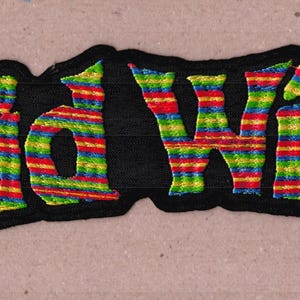 Könnte beinhalten: Ein gestickter Aufnäher mit dem Text Acid Witch in einer stilisierten Schriftart. Die Buchstaben sind mit horizontalen Streifen in leuchtenden Regenbogenfarben wie Rot, Gelb, Grün und Blau gefüllt und von einem dicken schwarzen Rand umgeben.