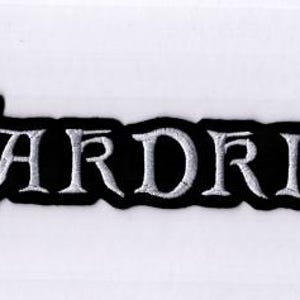 Puede incluir: Un parche bordado negro con la palabra "WARDRUNA" en blanco, con letras góticas. El parche tiene un borde negro y está sobre un fondo blanco. El texto es el elemento principal.