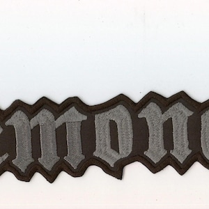 Pode incluir: Um emblema de tecido bordado com a palavra Demonaz numa fonte de inspiração gótica. As letras são feitas com fio cinzento claro e têm um contorno preto, criando um contraste nítido contra o fundo de cor branca.