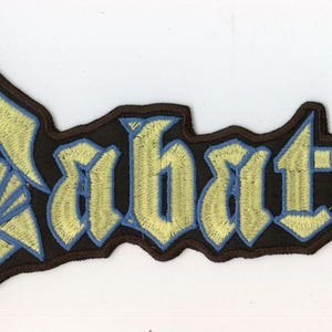 May include: An embroidered fabric patch featuring the word Sabaton in a stylised gothic font. The lettering is light yellow with a thin blue border, set against a black background with a dark brown stitched edge following the text contour.
