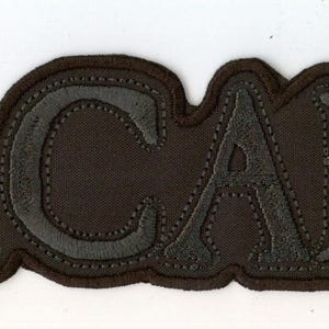 Pode incluir: Um emblema bordado em castanho escuro com a palavra MACABRE numa fonte serifada a negrito. As letras são contornadas com uma borda costurada em castanho escuro a condizer que segue o contorno do texto, criando um aspeto uniforme.