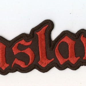 Pode incluir: Um emblema bordado retangular apresentando a palavra Enslaved em uma fonte gótica vermelha estilizada. As letras são contornadas com fio preto, criando um contraste ousado contra o fundo escuro. O emblema possui uma borda serrilhada distinta.