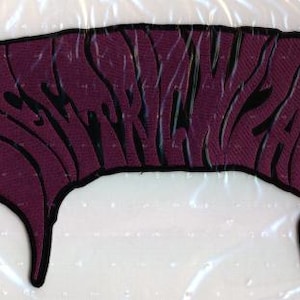 May include: A dark purple embroidered patch with a black border featuring the text ELECTRIC WIZARD in a stylized, distorted font. The letters are elongated and curved, creating a flowing, liquid-like appearance across the fabric surface.