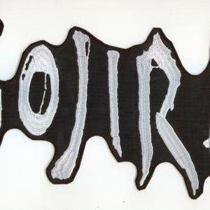 Könnte beinhalten: Schwarzer Aufnäher mit dem Wort "GOJIRA" in weißer, stilisierter Schrift. Der Aufnäher hat eine einzigartige, unregelmäßige Form mit schwarzem Hintergrund und weißen Nähten. Das Design ist auffällig und auffallend.
