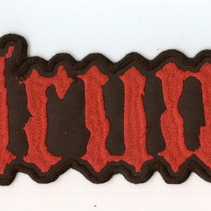 Könnte beinhalten: Ein rechteckiger gestickter Aufnäher mit dem Text "helrunar" in einer stilisierten, gezackten roten Schriftart. Die Buchstaben sind mit einem dicken schwarzen Rand versehen, der der Kontur der Zeichen folgt und einen Kontrast zum Rot bildet.