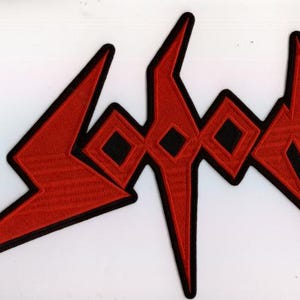 May include: Embroidered patch featuring the word "Sodom" in a stylized, angular design. The patch has a red background with black outlines and accents. The design includes sharp angles and geometric shapes.