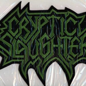 May include: A black and green embroidered patch featuring the text "CRYPTIC SLAUGHTER" in a stylized, angular font. The patch has a jagged, pointed border and is likely for clothing or accessories.