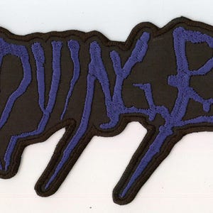 May include: A rectangular embroidered patch featuring the text MY DYING BRIDE in a stylized, jagged, dark purple font. The lettering is set against a black background with a thick black border that follows the contour of the letters.