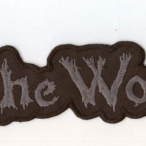 Op de afbeelding: Een zwarte stoffen patch met de tekst In The Woods... geborduurd in een lichtgrijs, getextureerd, takachtig lettertype. De patch heeft een gestanste rand die de onregelmatige vorm van de letters volgt en een duidelijk silhouet vormt.