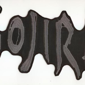 May include: Black patch with the word "GOJIRA" in gray, stylized lettering. The patch has a unique, irregular shape with a gray border. The design is simple yet bold, suitable for jackets, bags, or other fabric items.