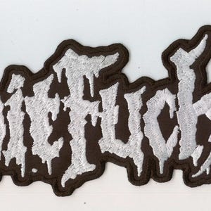 Könnte beinhalten: Ein bestickter Stoffaufnäher mit dem Wort Shitfucker in einer stilisierten, tropfenden silberweißen Schriftart mit einem dicken schwarzen Rand. Die Buchstaben haben eine texturierte, metallische Optik auf einem dunklen Hintergrund.