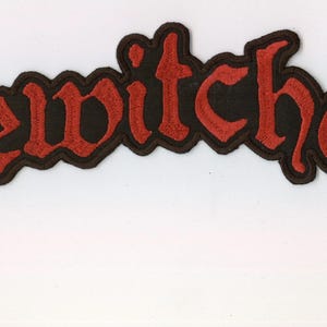 Könnte beinhalten: Ein gestickter Aufnäher mit dem Wort bewitcher in rotem Garn und schwarzem Rand. Die Buchstaben sind in einer bogenförmigen Anordnung gestaltet, wobei der erste und der letzte Buchstabe nach unten in kreuzartige Formen auslaufen.