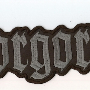 以下が含まれることがあります： 黒い布製のワッペンで、グレーのゴシック体で「Gorgoroth」という文字が刺繍されています。ワッペンには文字の輪郭に沿った太いステッチの縁取りがあり、暗い背景に対してはっきりとしたアウトラインを描いています。