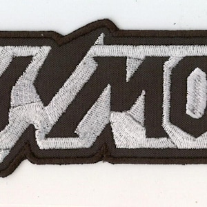 May include: A rectangular embroidered patch featuring the text HOLY MOSES in a bold, stylised black font. The letters are set against a light grey, textured background with a thick black border that follows the angular shape of the lettering.