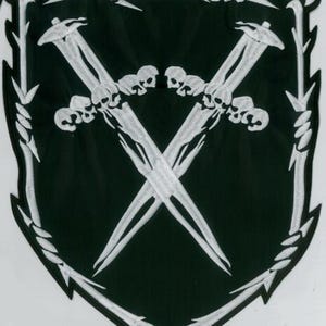 May include: A black and white embroidered patch featuring a shield design. Two crossed swords with skull details are centered on the patch. The border is white with a barbed wire and leaf design.