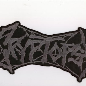 May include: A black patch with gray embroidered text that reads "SATANAS". The patch has a jagged, irregular border. The text is in a gothic, stylized font, and the patch is likely for clothing or accessories.