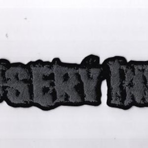 May include: A black and gray embroidered patch with the text "MISERY INDEX" in a stylized font. The patch has a black border and is likely designed to be sewn or ironed onto fabric. The letters are in a textured, distressed style.